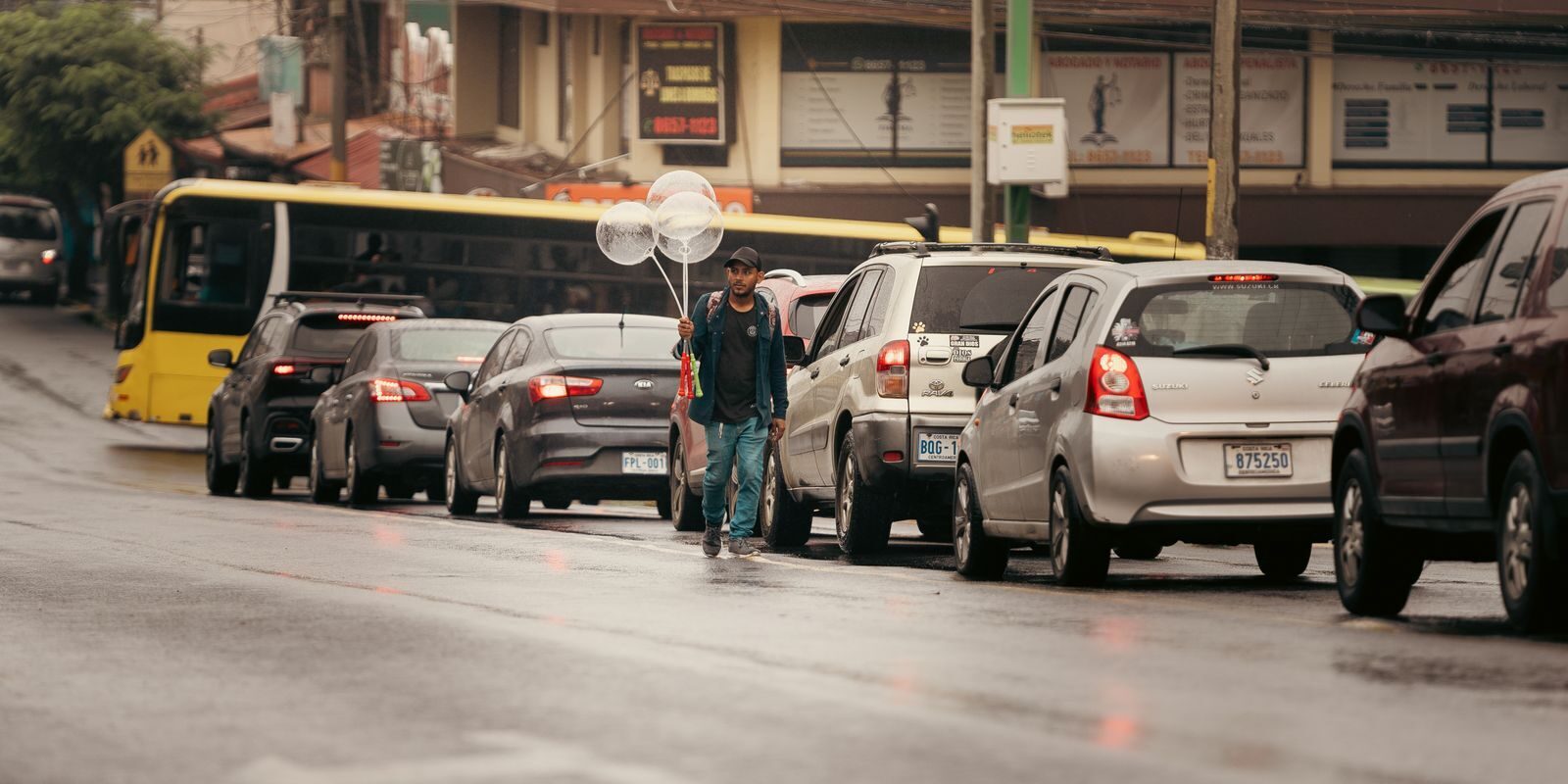 A street vendor in Alajuela, Costa Rica, selling balloons amidst traffic. Captured on a rainy day.