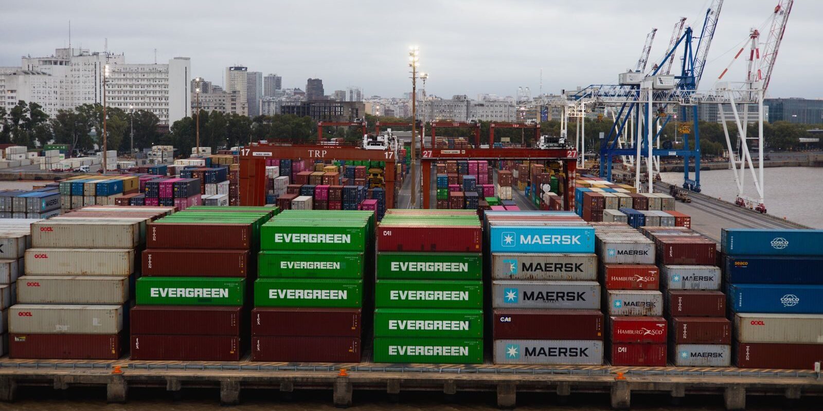 Aerial view of colorful shipping containers in a bustling port with cranes and city skyline.