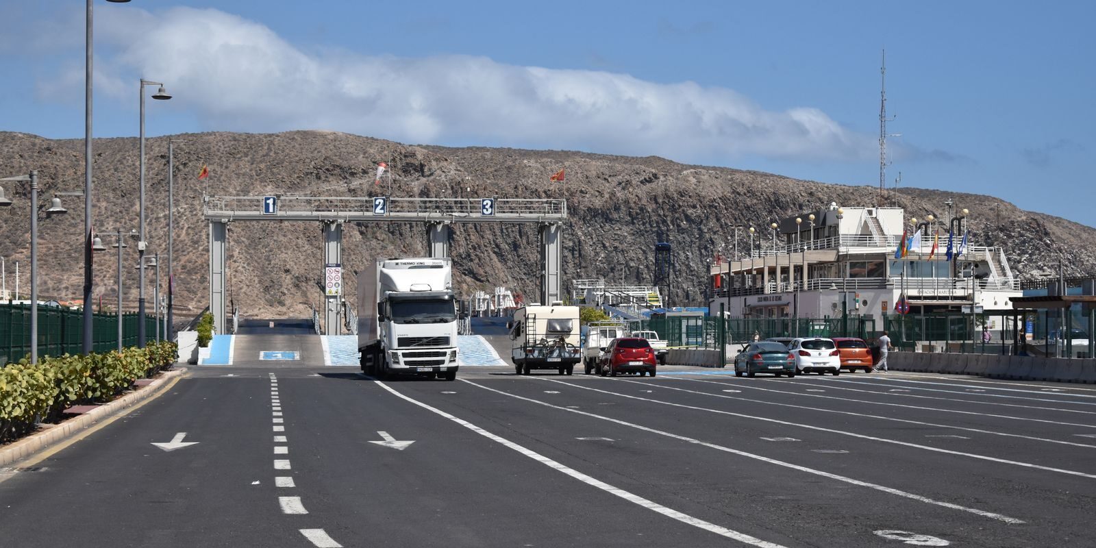A ferry terminal with trucks and cars under a clear blue sky and dry hills in the background.