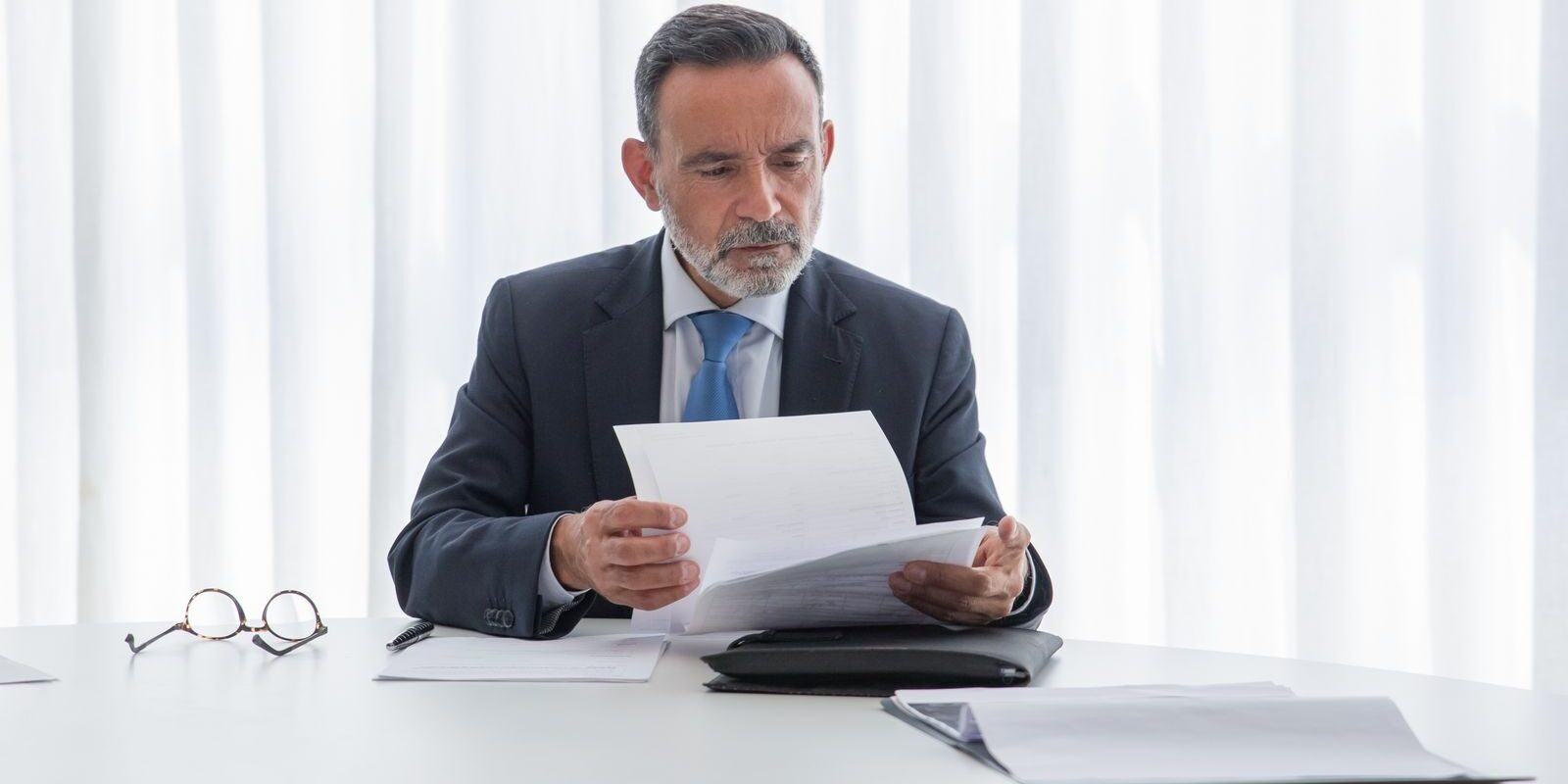 Photo by Kampus Production Elderly businessman focusing on reading documents at a table in a bright office setting.